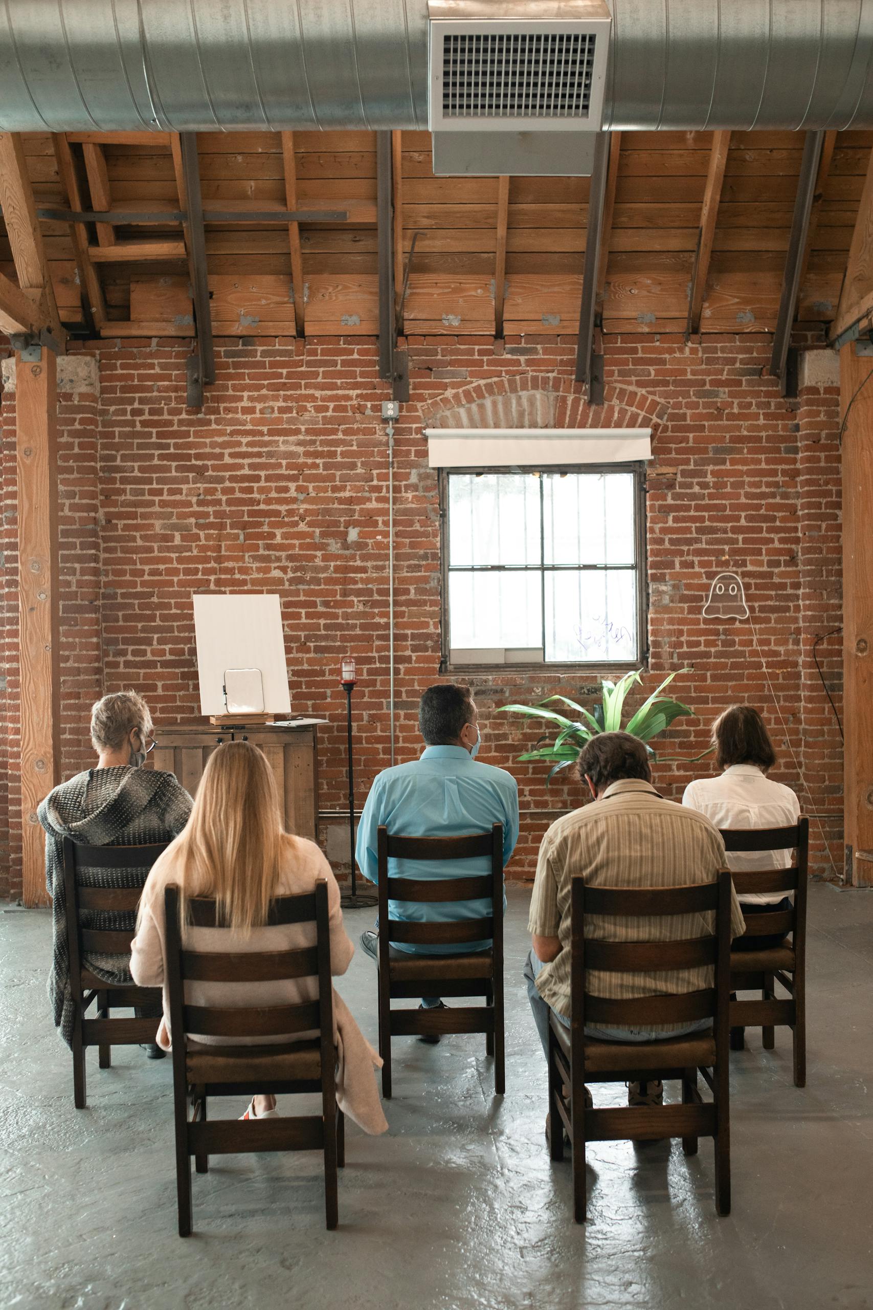 A group of people sitting indoors facing an easel in a rustic brick-walled room.