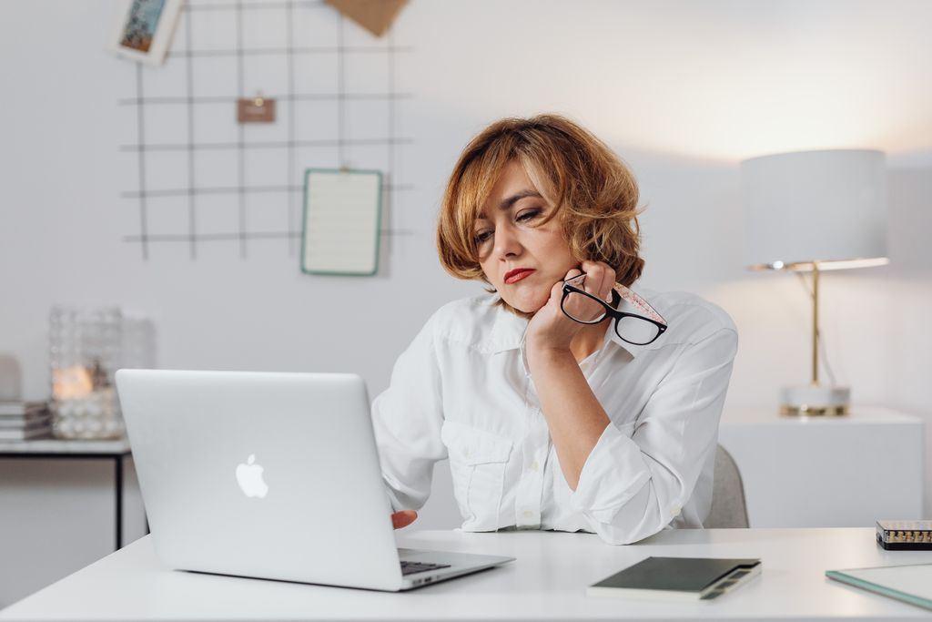 Woman in white shirt working on laptop at home office desk with eyeglasses in hand.